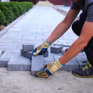 Un homme en gants de travail installe des pavés gris sur un lit de sable, créant une allée. Travaux d'aménagement paysager.