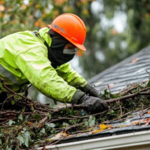 Travailleur en tenue de pluie orange et jaune nettoyant des branches et feuilles d'une gouttière sur un toit mouillé.